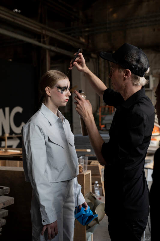 Mustapukuinen meikkaaja levittää dramaattista silmämeikkiä airbrushilla valkoiseen asuun pukeutuneelle mallille, joka seisoo kampaamo Helsingin teollisuustyylisessä backstage-tilassa.