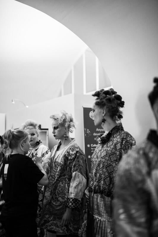 Mustavalkoinen valokuva useista ihmisistä, joilla on hienostunut kampaus ja rohkea meikki ja joilla on kuvioituja asuja, ja jotka seisovat sisätiloissa. Yksi t-paitainen henkilö, ehkä helsinkiläisestä kampaamosta, näyttää avustavan ryhmää.