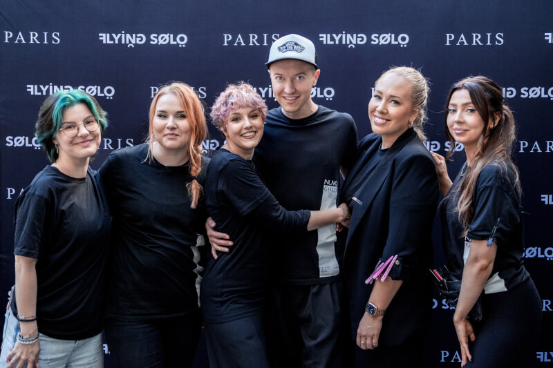 Kuusi ihmistä seisoo hymyillen yhdessä mustan taustan edessä, jossa on "Flying Solo" ja "Paris" -logot. Heidän tyylikkäät ulkonäkönsä kääntäisivät katseita missä tahansa Helsingin kampaamossa, sillä raikkaat hiusten raidotus ja tyylikkäät mustat asut luovat juhlavaa tunnelmaa.