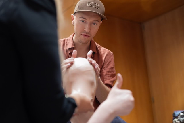 Kampaamo Helsingissä ruskeaan lippikseen ja paitaan pukeutunut henkilö säätää varovasti mallinuken päätä, kun toinen nostaa peukkua. Viihtyisässä sisätilassa on puiset seinät.