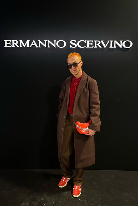Henkilö, jolla on aurinkolasit, pitkä ruskea takki, punainen paita, ruskeat housut ja punaiset lenkkarit, pitelee poseeraa mustan seinän edessä, jossa on ERMANNO SCERVINO -logo, ja hänen hiuksensa on muotoiltu täydelliseksi hiusten raidotuksella.